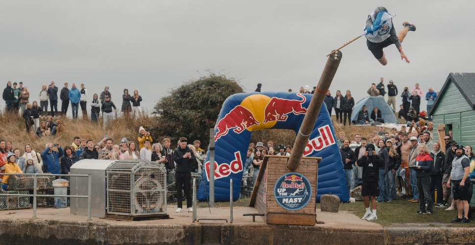 : A person wearing a helmet and a white shirt with a logo is airborne, holding a blue and white flag while participating in an outdoor event involving balancing on a large wooden pole. A crowd of spectators watches in the background, with an inflatable Red Bull archway visible on the left side.